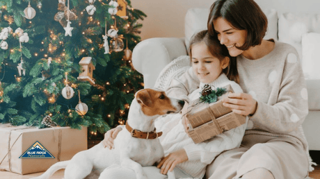 Mother holding her daughter, who is petting a Jack Russell Terrier with a Christmas tree in the background.