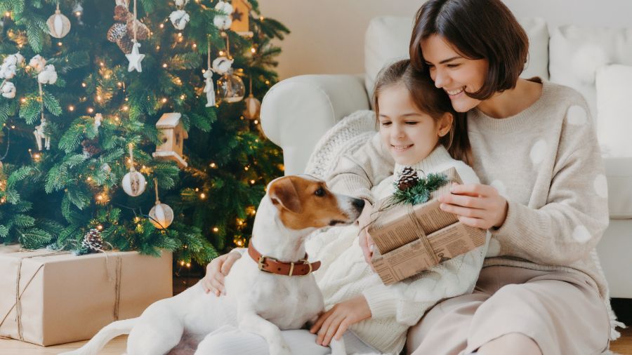Mother holding her daughter, who is petting a Jack Russell Terrier with a Christmas tree in the background.